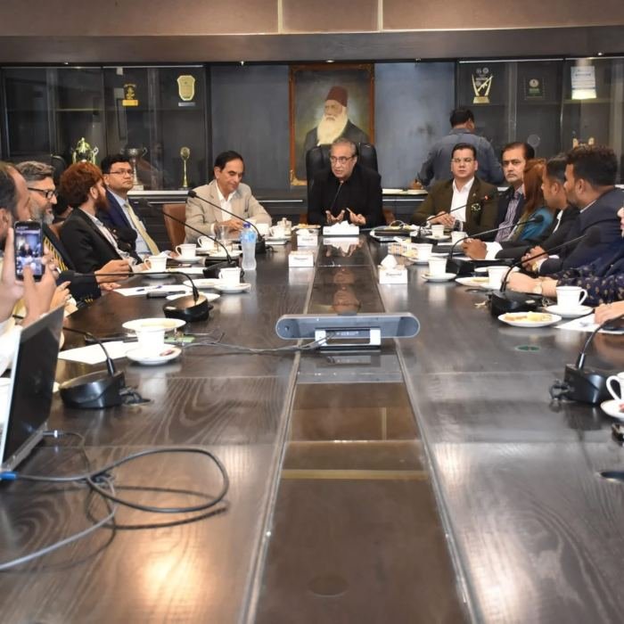 Delegates seated around a large wooden boardroom table with microphones and tea service, beneath a portrait of Sir Syed Ahmed Khan
