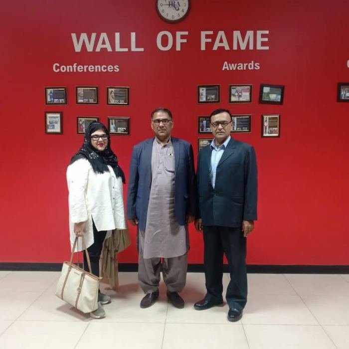 Three professionals standing in front of a red “Wall of Fame” display of framed conference and award photographs