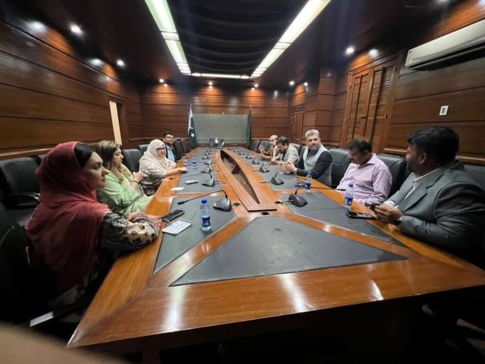 A small group in discussion around a wide committee table in a wood-panelled Sindh Assembly room, beside the national and provincial flags