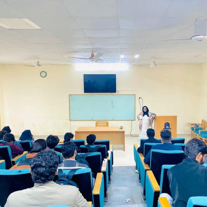 A speaker addressing rows of seated students from the front of a university lecture hall
